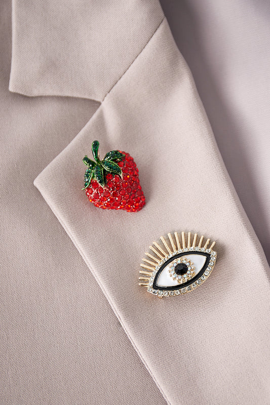 Close-up of beige blazer lapel decorated with a red rhinestone strawberry brooch and gold-tone eye brooch with rhinestones