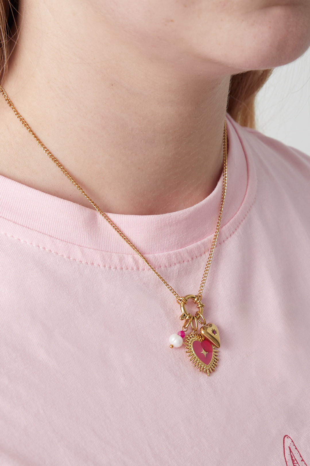 Gold pendant necklace with red enamel heart, pearl and pink flower charms worn over pink t-shirt