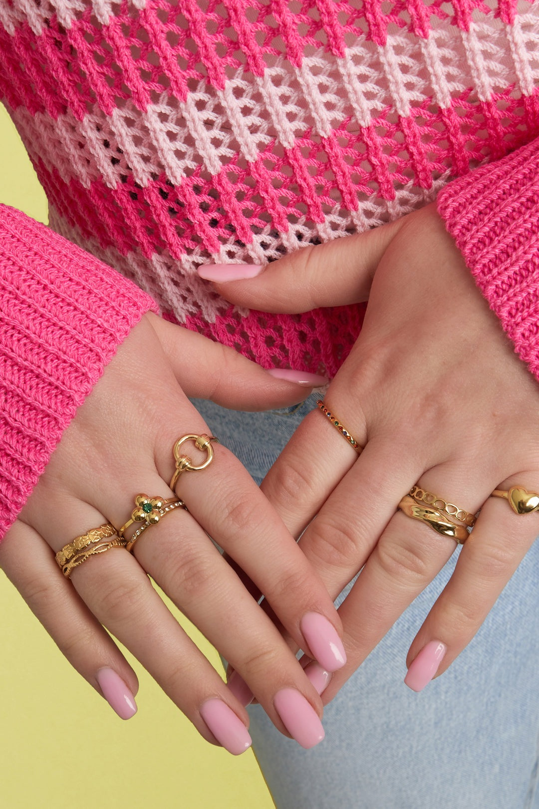 Woman's hands adorned with multiple gold rings, including a braided gold minimalist ring, wearing pink knit top and light blue jeans