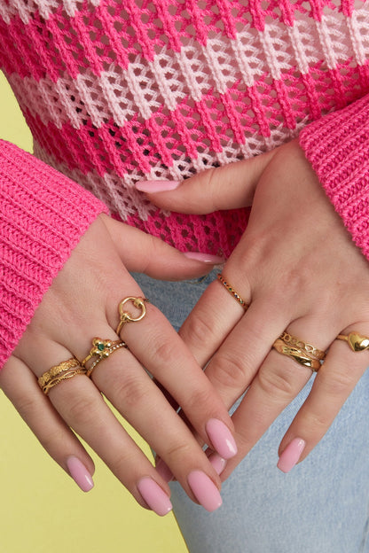 Woman's hands adorned with multiple gold rings, including a braided gold minimalist ring, wearing pink knit top and light blue jeans