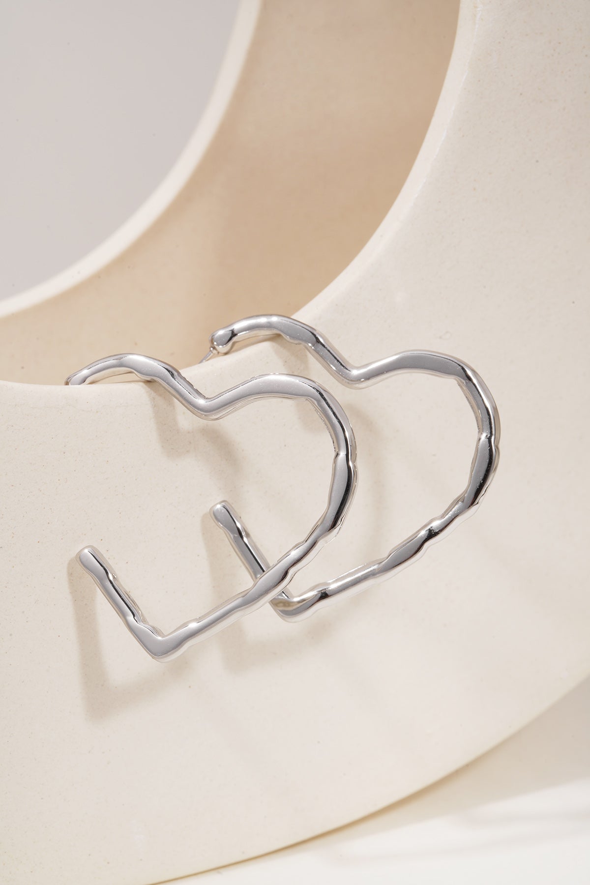 Silver-tone heart-shaped hoop earrings with a textured, molten metal finish displayed on a curved ceramic stand.