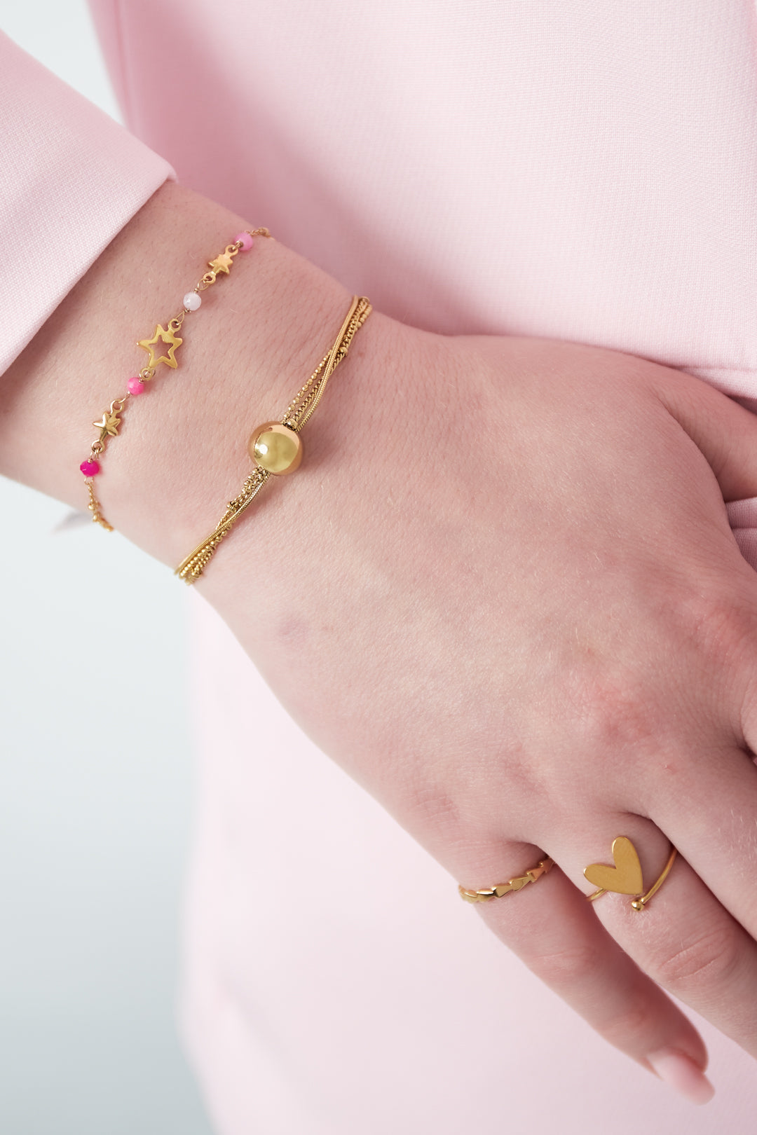 Model wearing a gold multi-chain sphere charm bracelet paired with a pink star-accented bracelet and gold heart ring on wrist