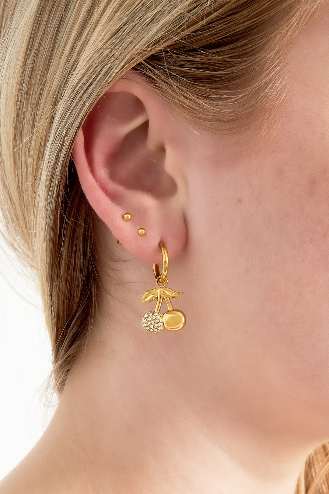 Close-up of a woman wearing one gold cherry drop earring with rhinestone detail, paired with two small gold stud earrings
