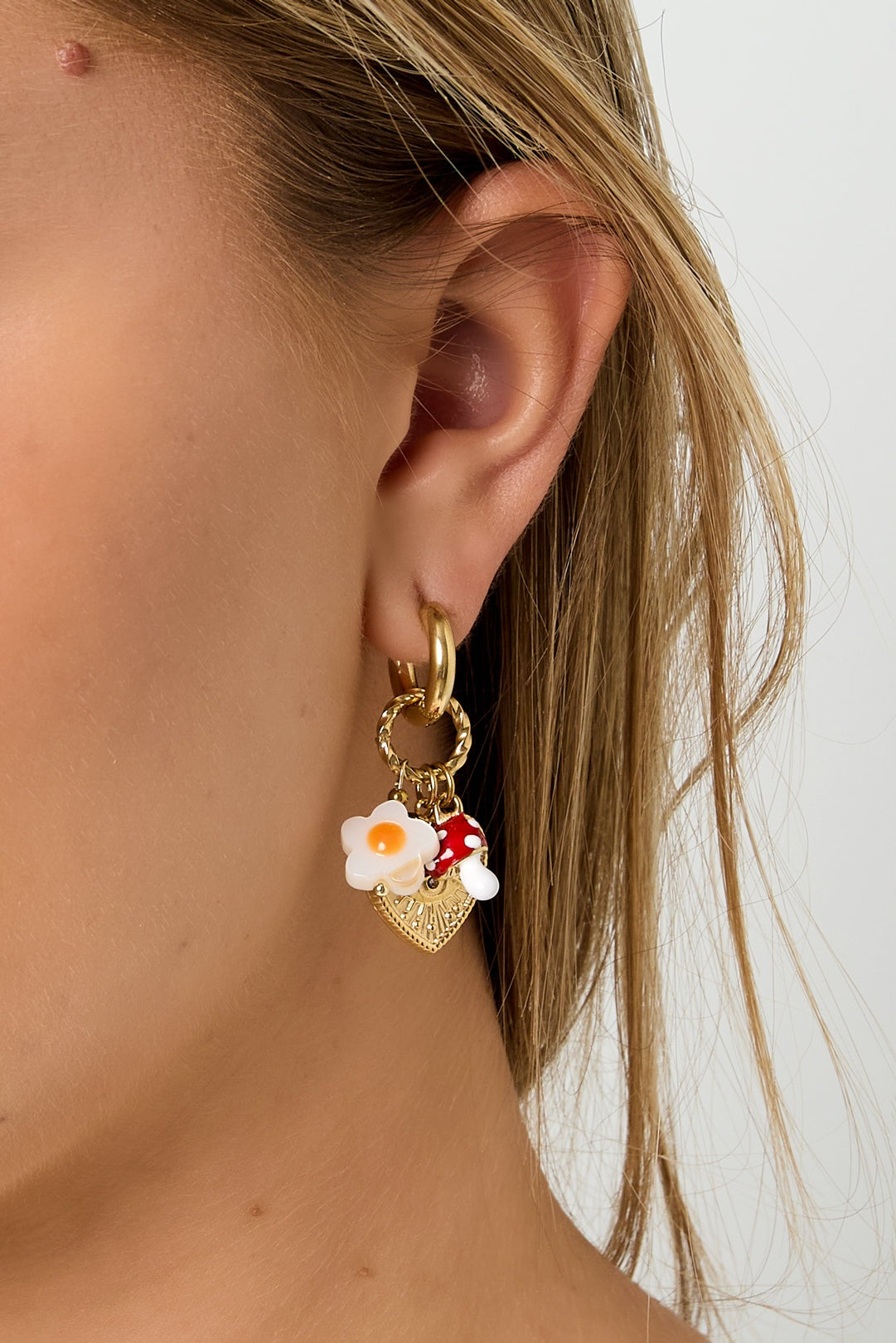 Side profile of woman wearing gold charm hoop earring showing mushroom, flower, and heart details against blonde hair