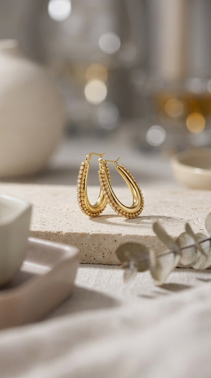 Drip Drop Glow earrings: gold teardrop hoops with beaded detailing and pave crystal accents on a stone surface.