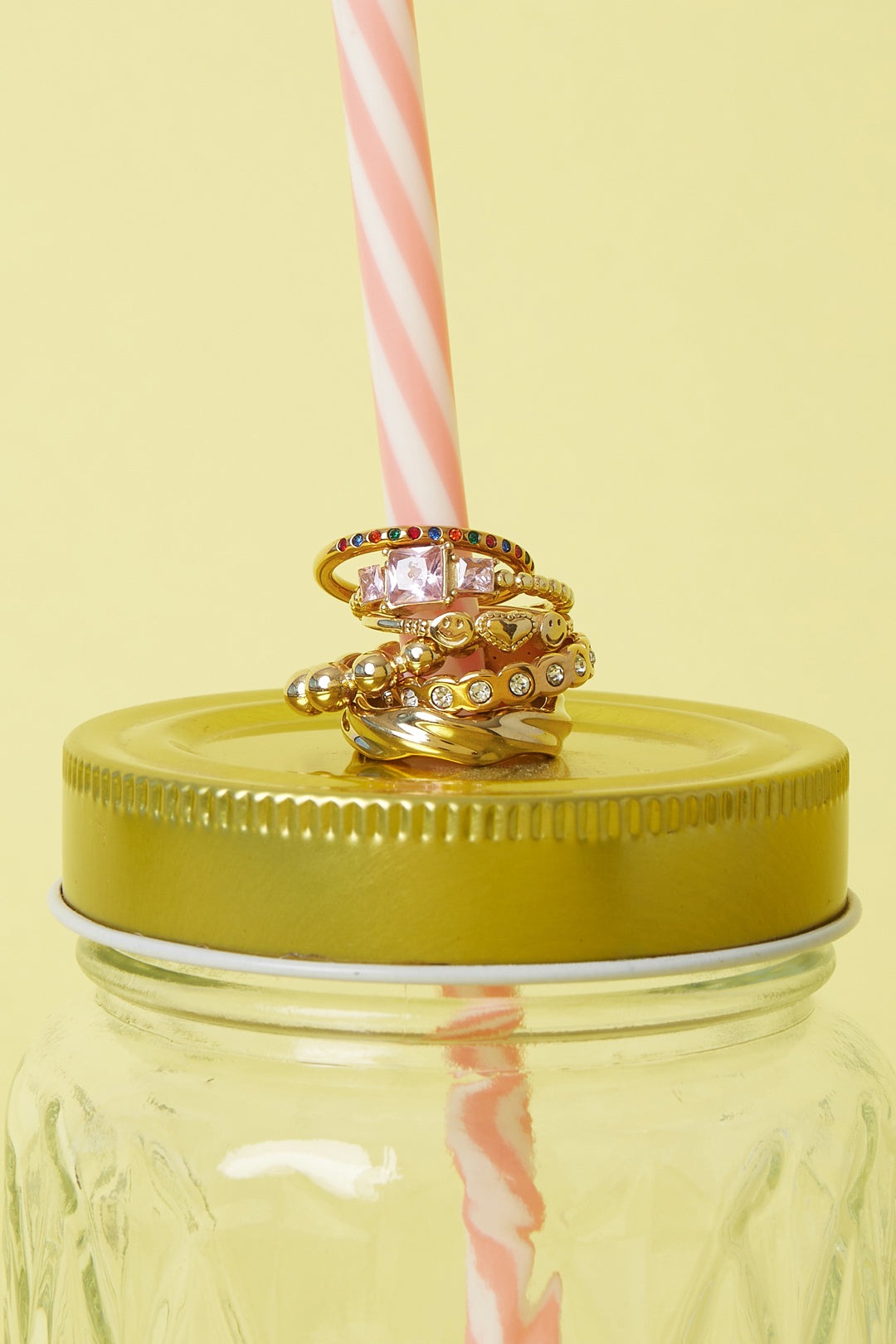 Stack of diverse gold rings including one with pink square gemstones placed on a jar lid with a striped straw