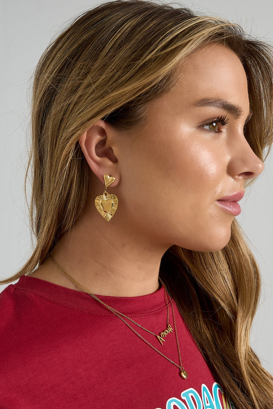Side profile of woman wearing gold-tone heart drop earrings paired with red top and gold necklaces