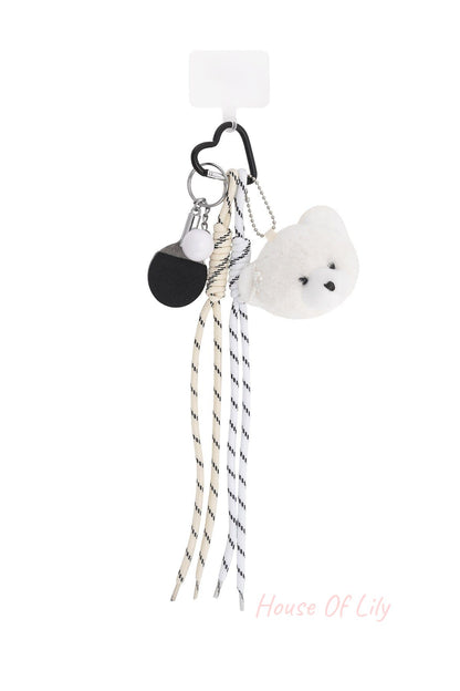 Plush bear bag charm featuring a white teddy bear head with pearl detail, striped fabric laces, mini ping pong paddle keychain, and heart-shaped black clip on a white background.