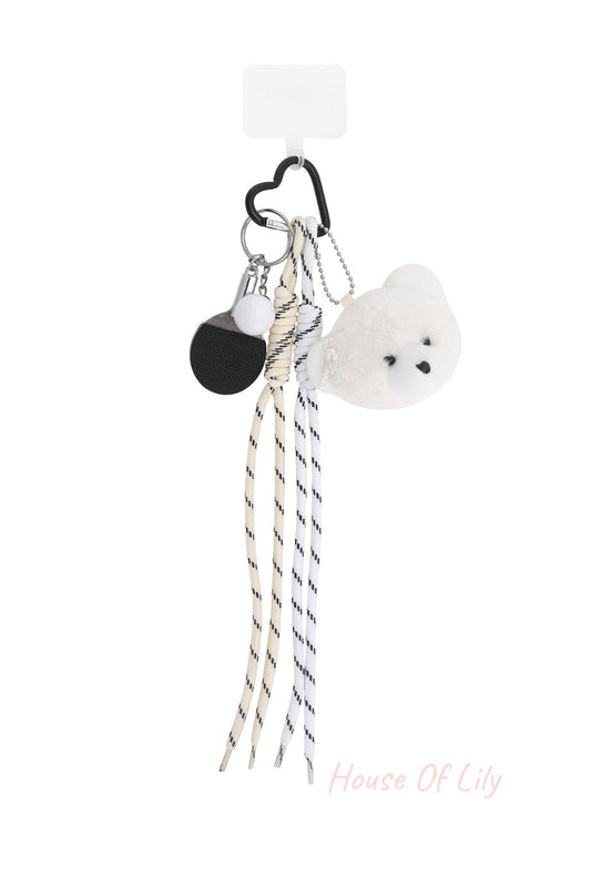 Plush bear bag charm featuring a white teddy bear head with pearl detail, striped fabric laces, mini ping pong paddle keychain, and heart-shaped black clip on a white background.