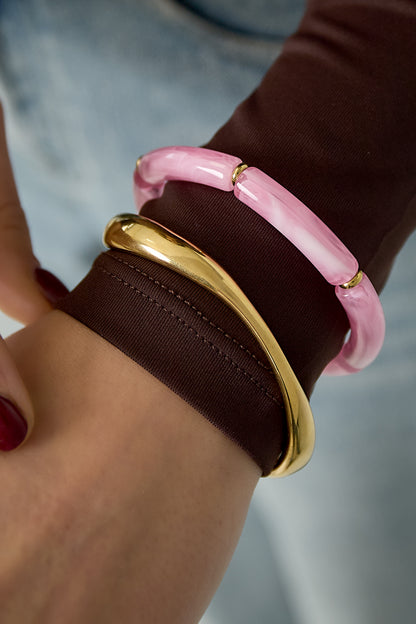 Close-up of wrist wearing a light pink resin tube bracelet and a shiny gold bangle over brown clothing