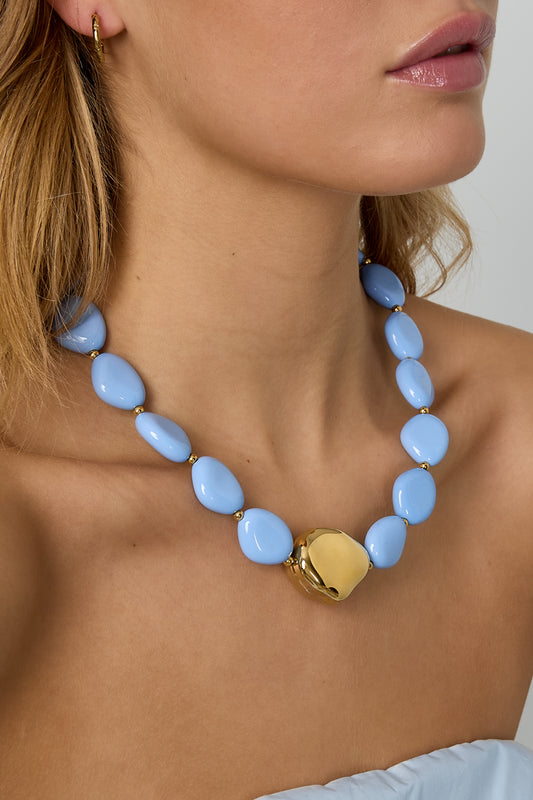 Close-up detail of model's neck showing glossy blue irregular beads and gold-tone centerpiece on necklace