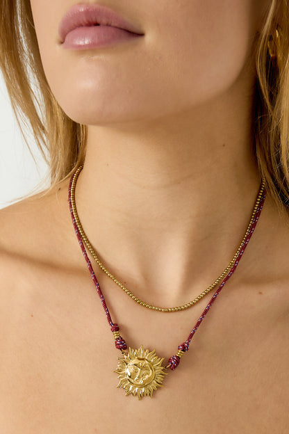 Close-up of model's neck showcasing layered gold-tone sun pendant necklace on red rope with a gold beaded necklace