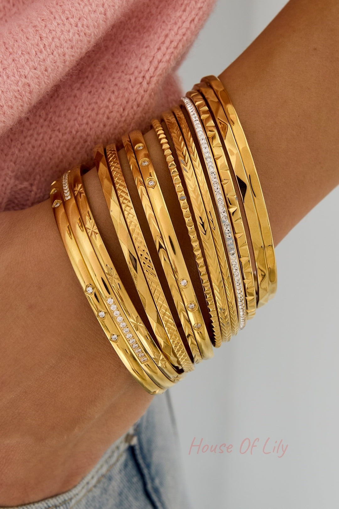 Wrist wearing a wide stack of various gold bangle bracelets with different textures and sparkle, combined with casual pink and denim outfit