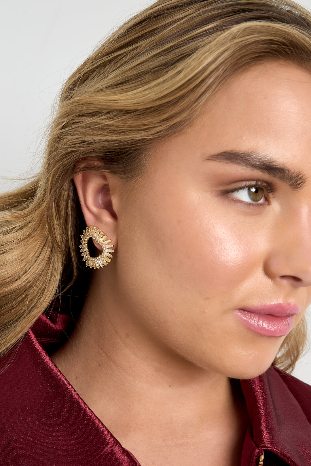 Model wearing gold-tone radiant heart stud earring, styled with soft waves and maroon satin top