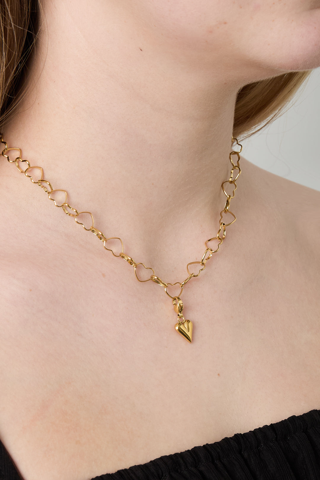 Close-up of model wearing gold heart link chain necklace featuring a small hanging heart charm, styled with black outfit