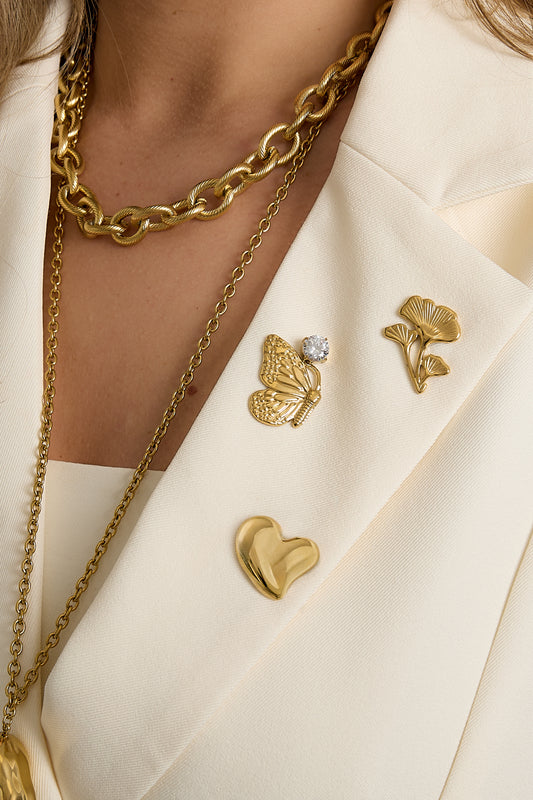 Woman wearing cream blazer adorned with three gold brooches: butterfly with rhinestone, floral design, and heart, paired with gold chain necklaces.