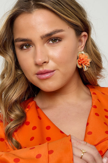 Close-up of woman wearing large orange floral raffia stud earring with gold-tone border, matching orange polka dot blouse