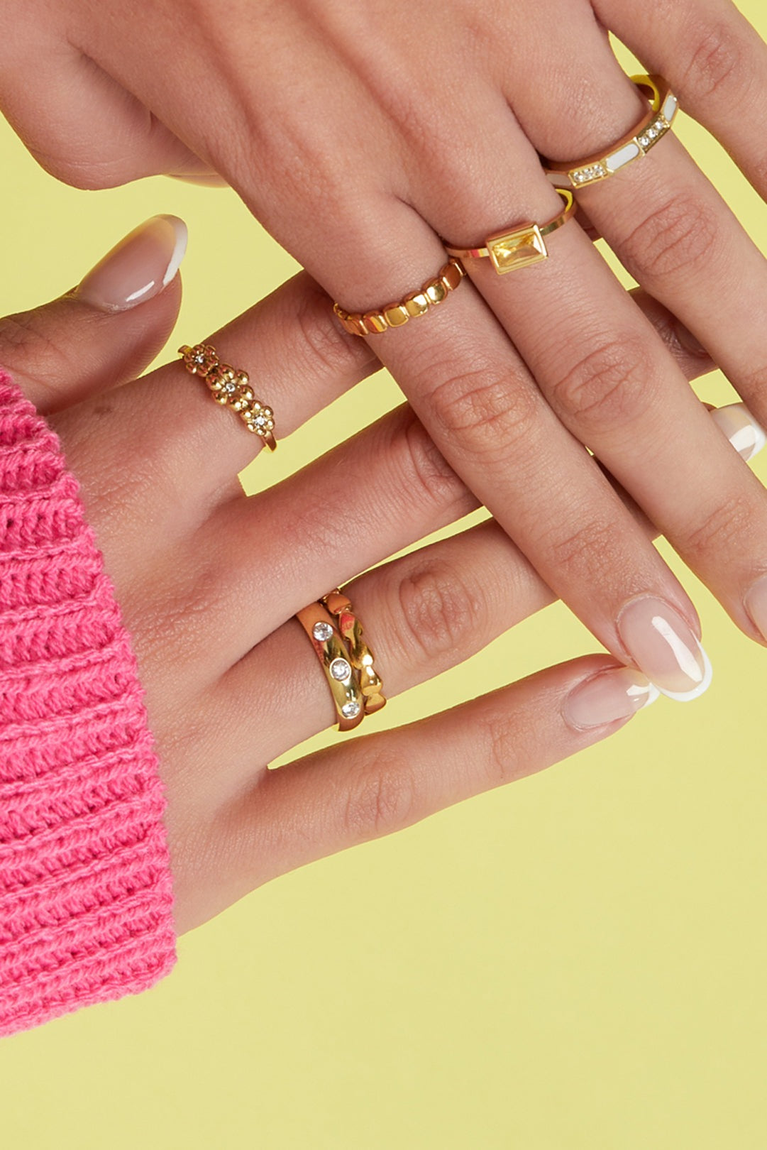 Close-up image of a woman's hand wearing multiple gold rings including a heart link ring on yellow background with pink sweater sleeve