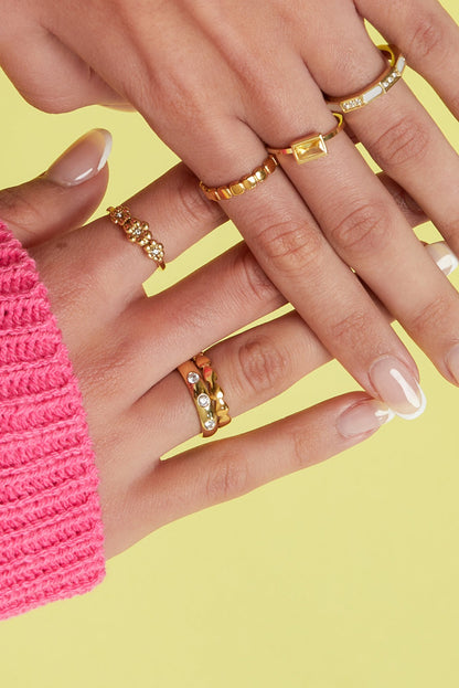 Close-up image of a woman's hand wearing multiple gold rings including a heart link ring on yellow background with pink sweater sleeve