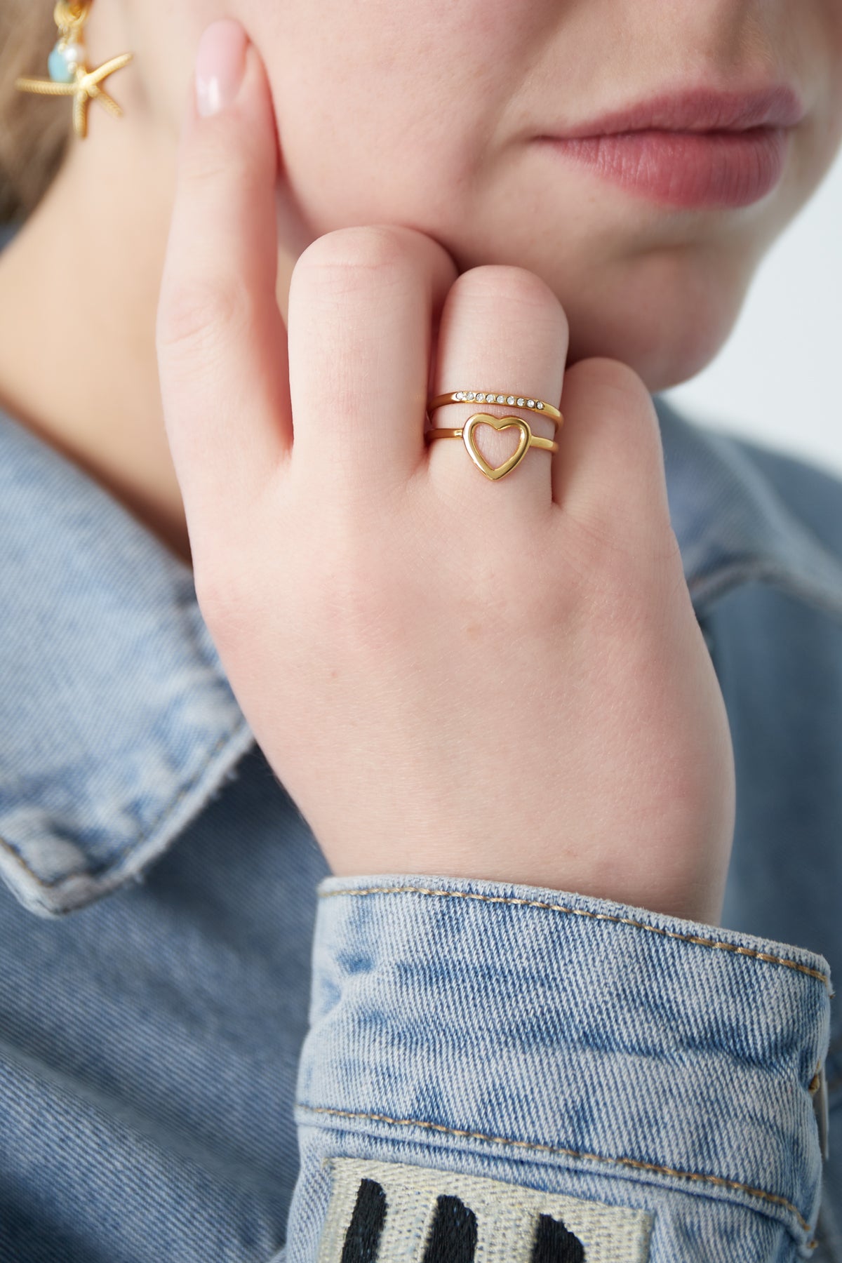 Close-up of a woman's hand wearing the gold heart accent ring with rhinestone band, paired with a denim jacket and a starfish earring