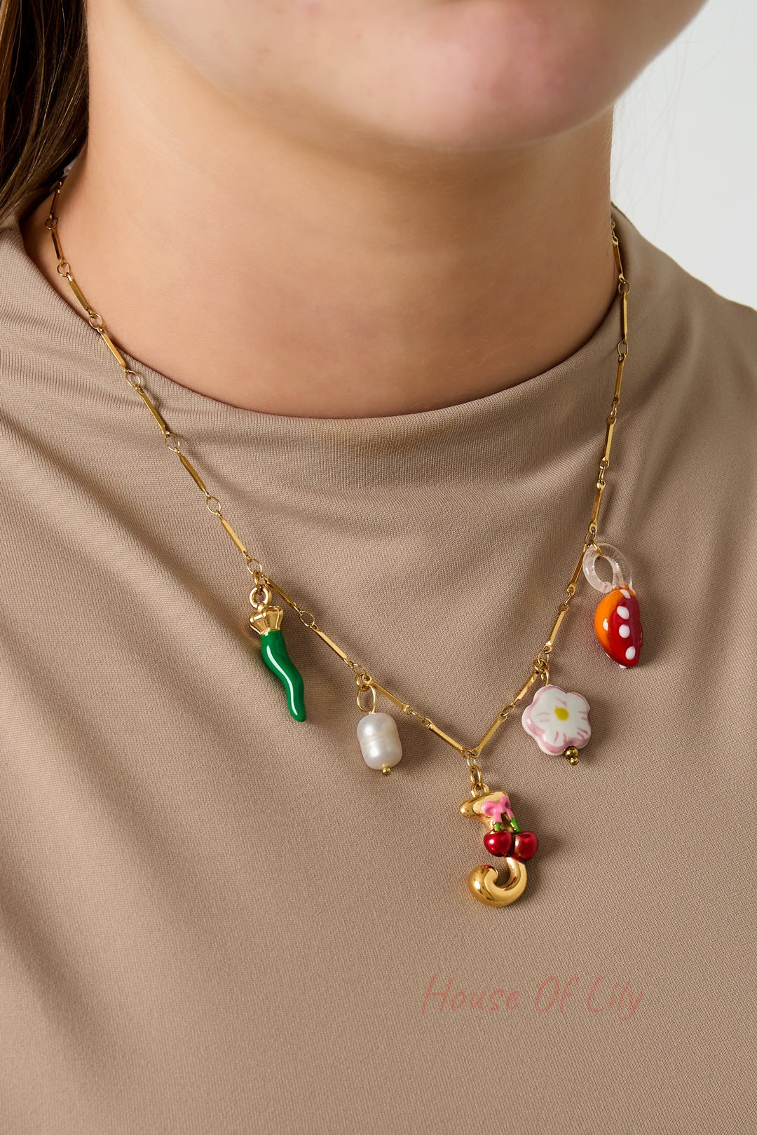 Model wearing a gold charm necklace with green chili pepper, white pearl, pink flower, red mushroom, and gold letter J with cherry pendant