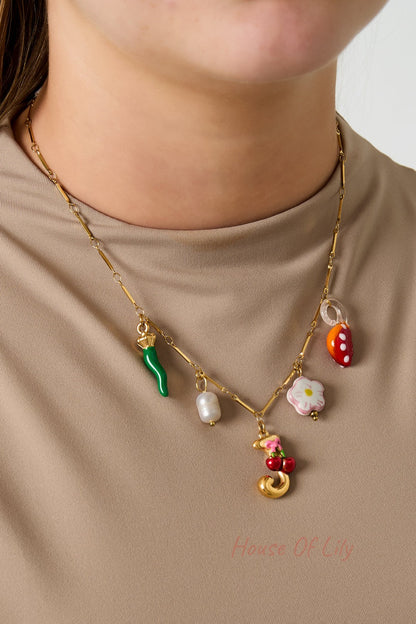 Model wearing a gold charm necklace with green chili pepper, white pearl, pink flower, red mushroom, and gold letter J with cherry pendant