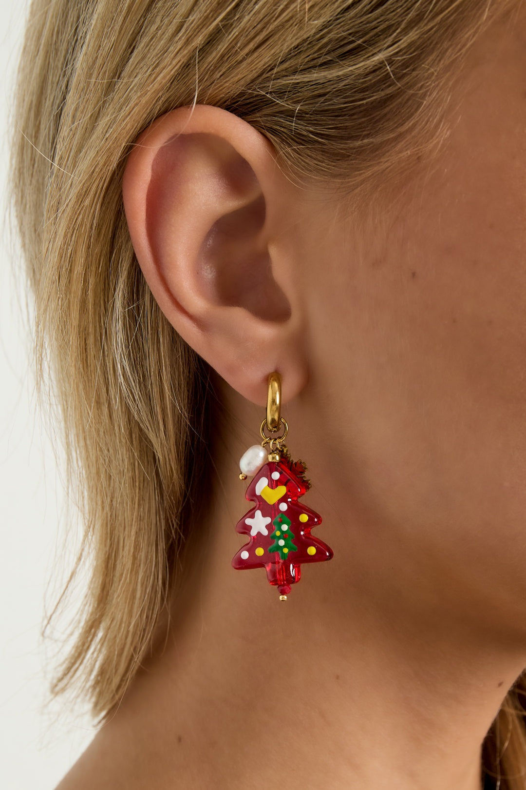 Close-up of a woman wearing a festive Christmas tree drop earring with gold hoop, red resin tree charm, gold snowflake, and white bead, showing how the earring hangs