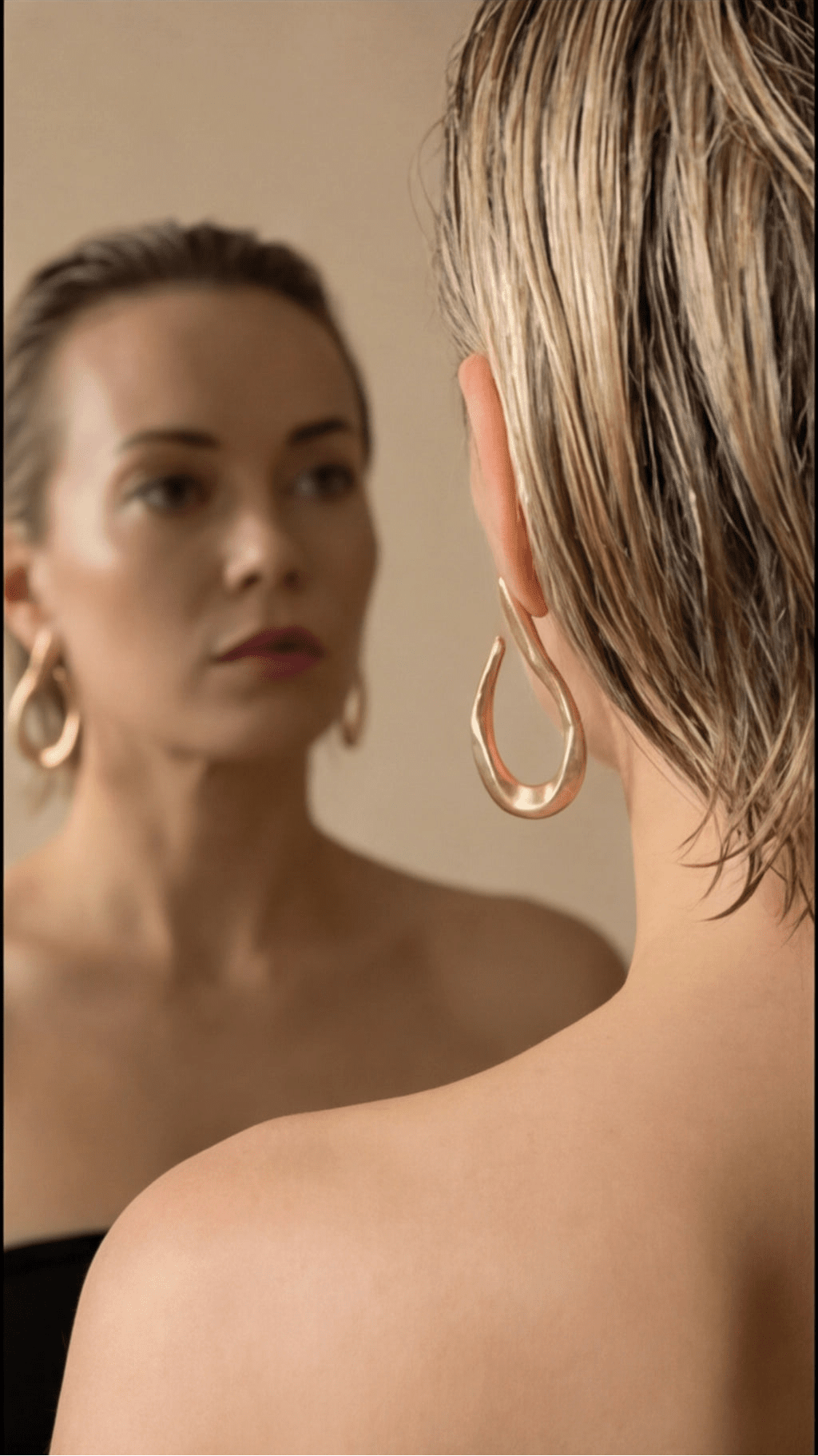 Woman wearing gold hoop earrings looking at herself in a mirror.