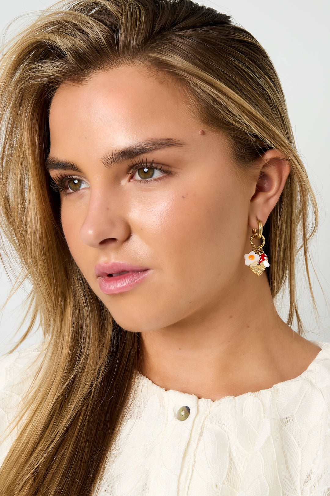 Close-up portrait of woman wearing gold hoop earrings with mushroom, flower, and heart charms, styled with a cream textured top