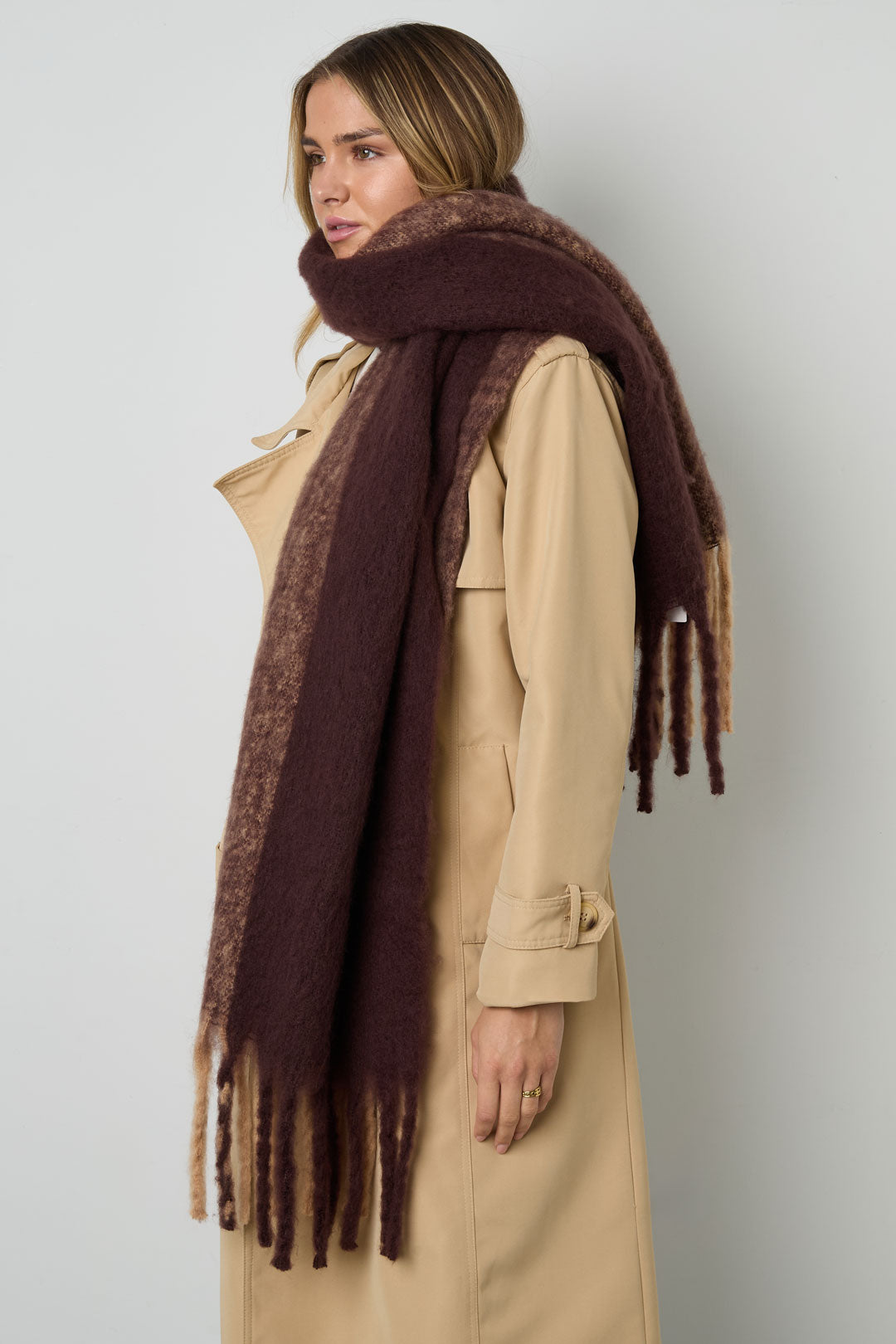 Model wearing a brown gradient fringe scarf styled over a beige trench coat, side view