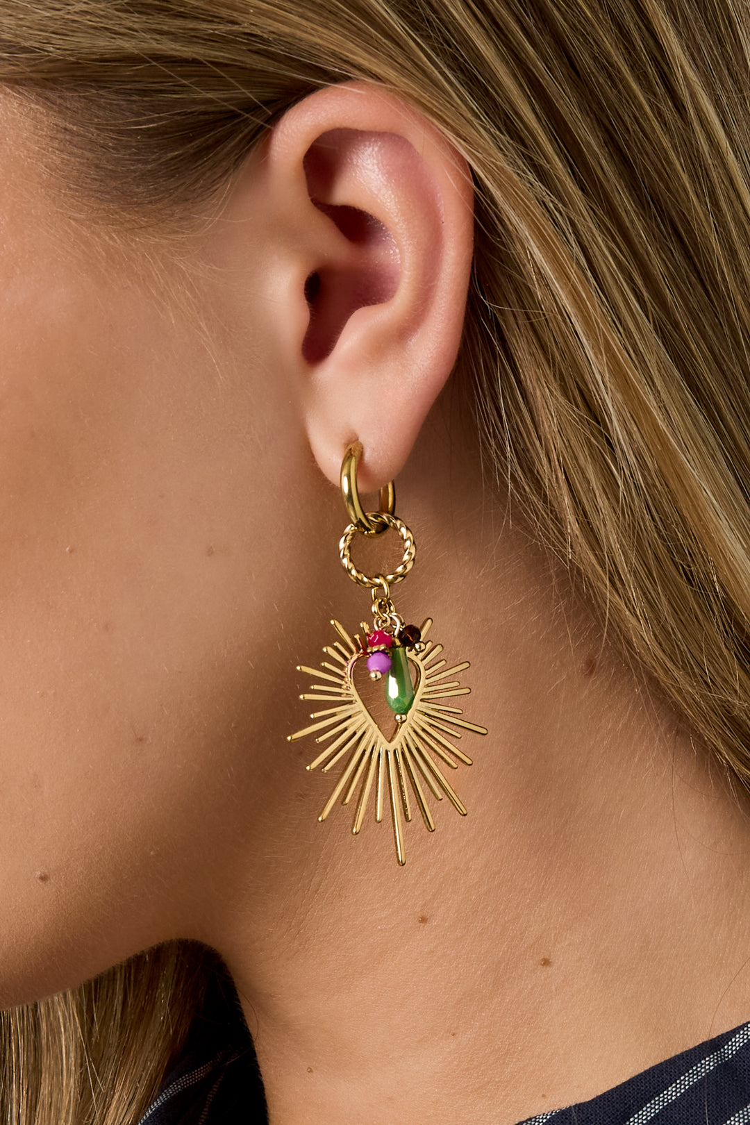 Close-up of woman’s ear wearing radiant heart charm gold sunburst drop earring with colorful bead accents