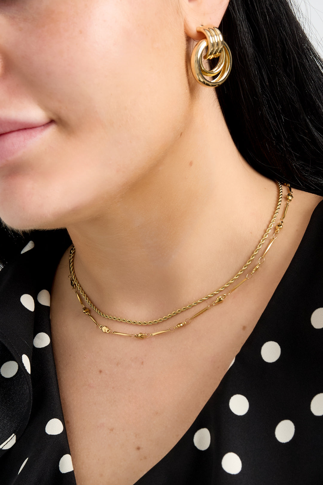 Model wearing layered gold necklaces including the heart link necklace paired with gold hoop earrings and a black polka dot top