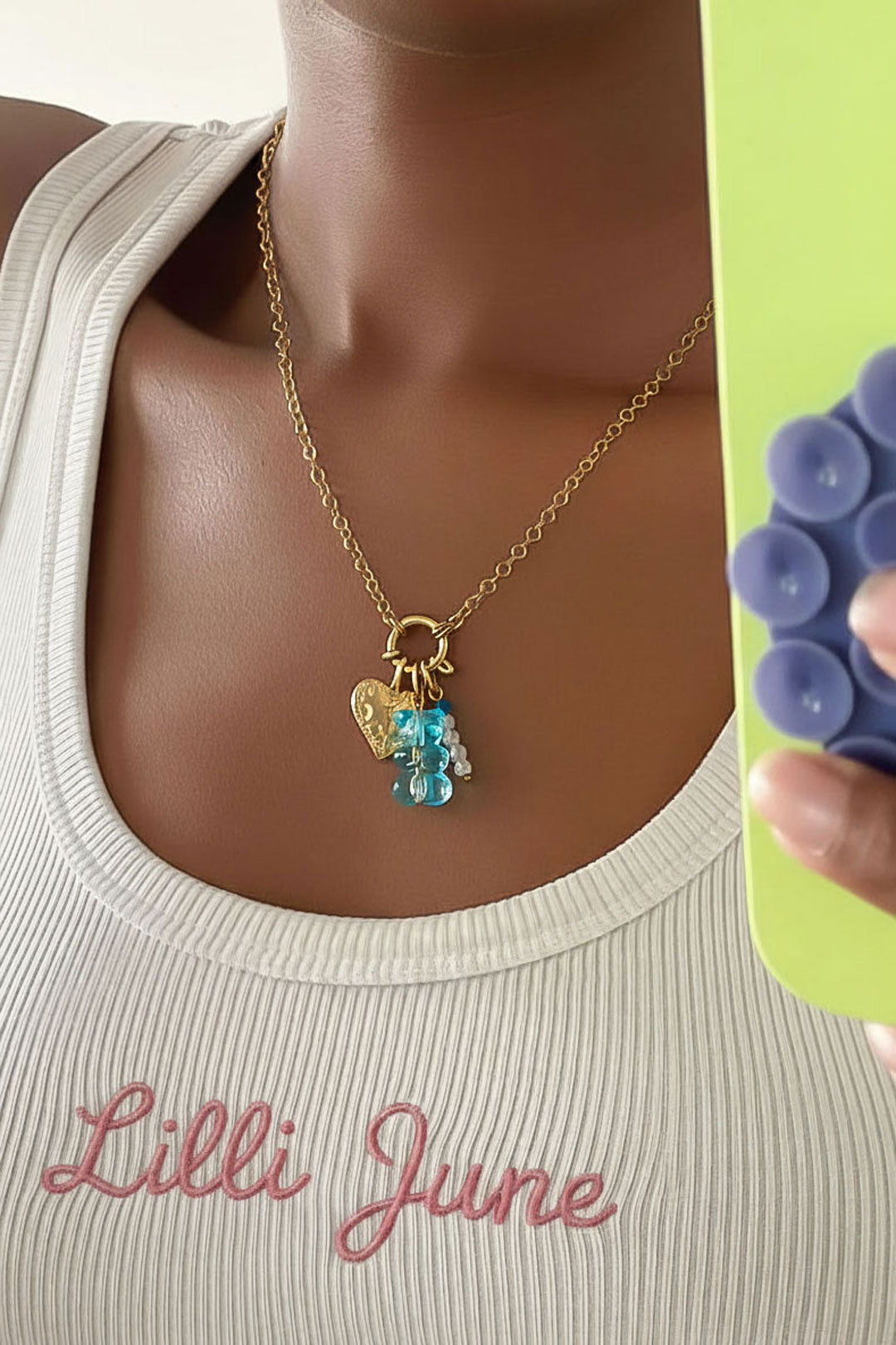 Close-up of necklace worn on woman with deep skin tone wearing a white ribbed top, showing blue gummy bear charm and gold heart pendant