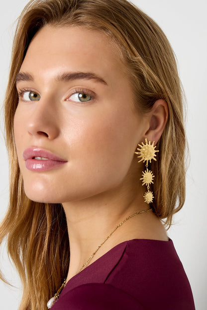 Side profile of woman wearing gold sunburst dangle earrings with maroon top