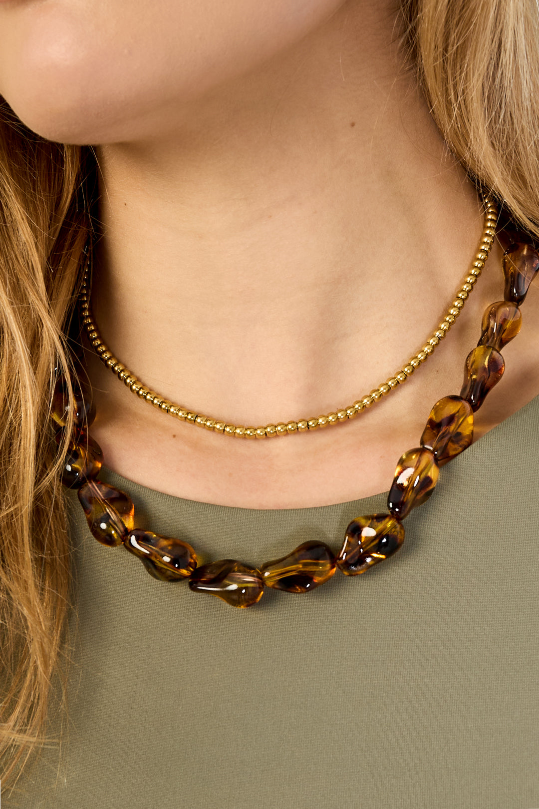 Close-up view of tortoiseshell beaded necklace and gold ball chain layered on model wearing olive top