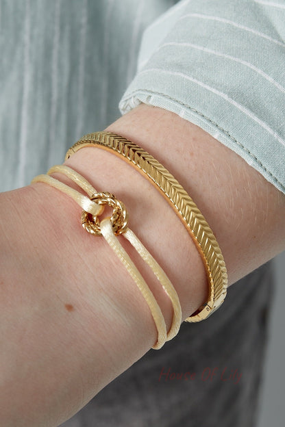 Close-up of a female wrist wearing a textured gold bangle bracelet and a cream-colored cord bracelet, paired with a light blue striped shirt