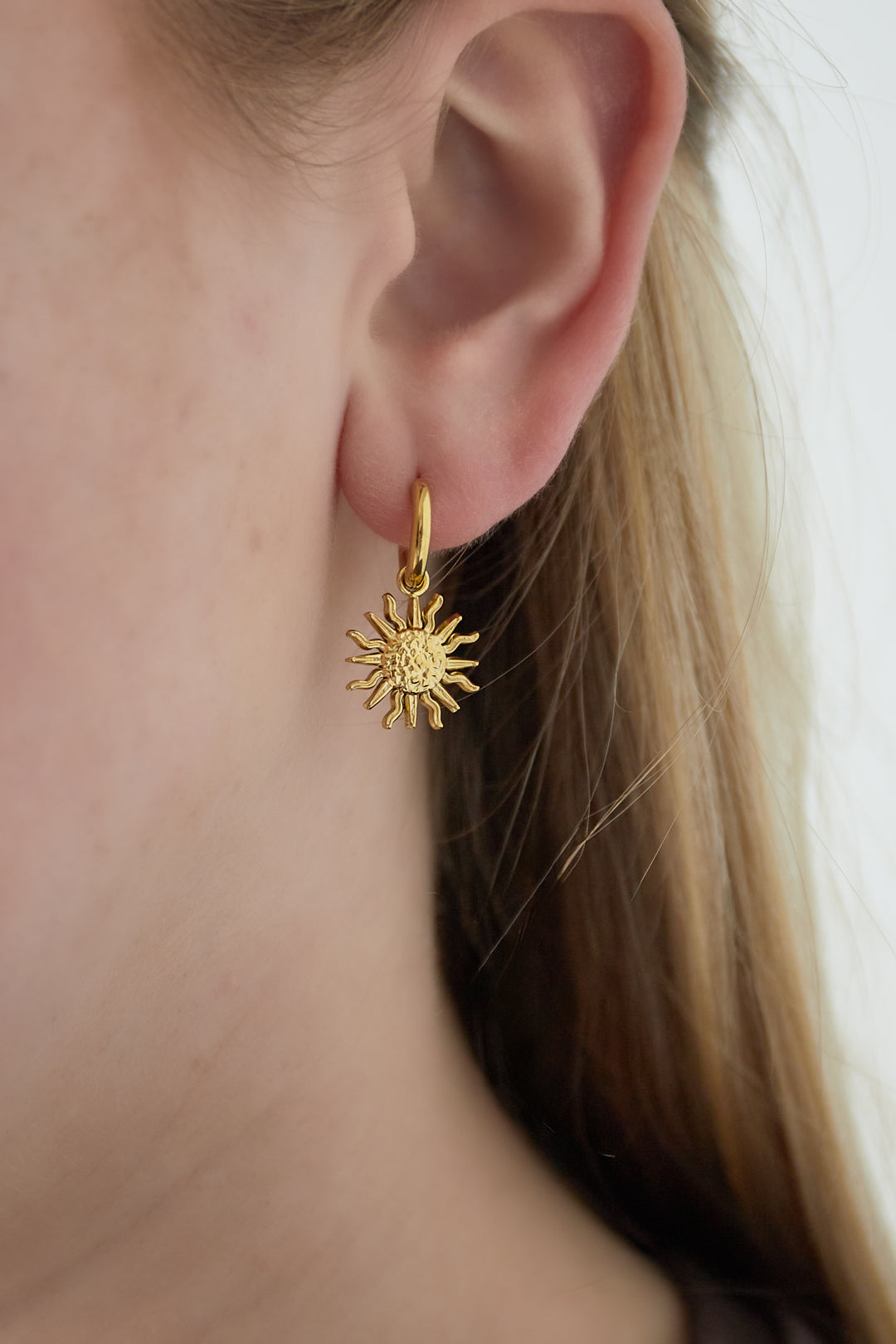 Close-up of gold sun charm hoop earring worn on a woman's ear against a light background