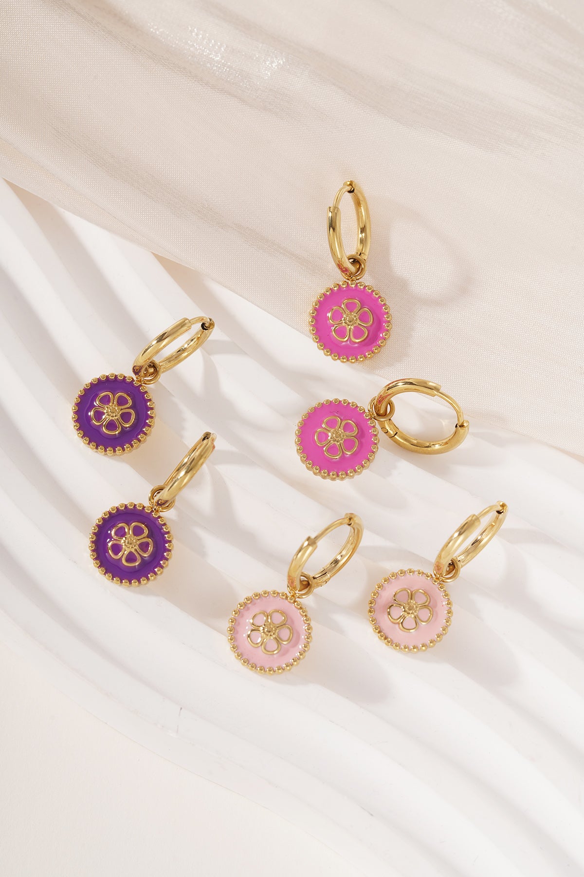 Display of multiple gold-tone hoop drop flower earrings in pink, purple, and rose enamel on white textured surface