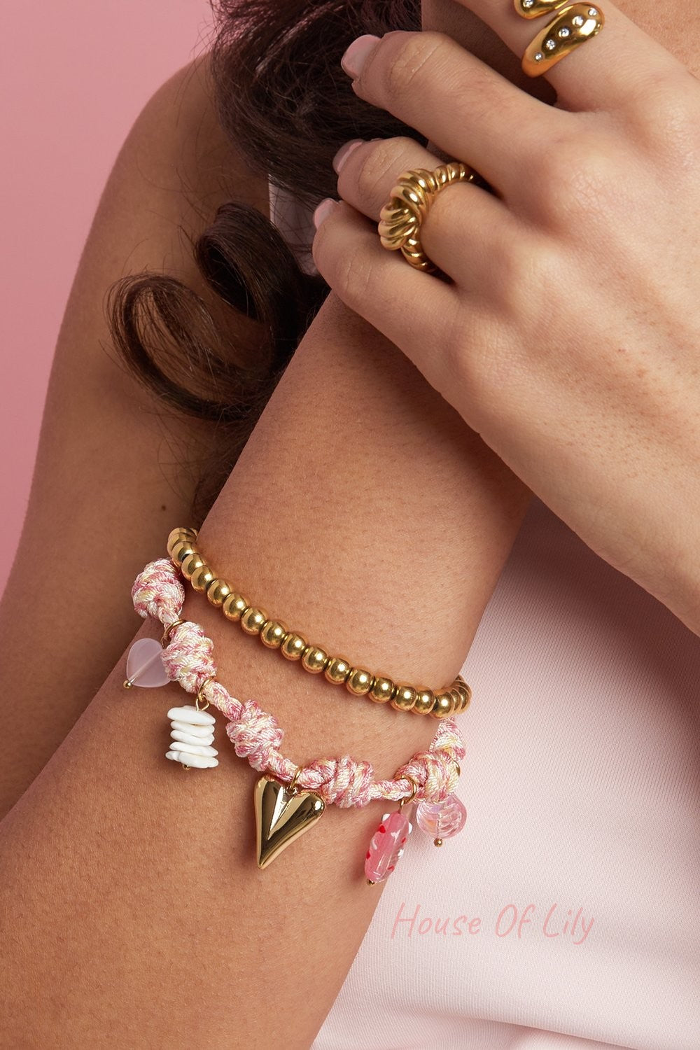 Nautical knot charm bracelet in pink cord with gold heart, shell, and bead charms, stacked with a gold beaded bracelet.
