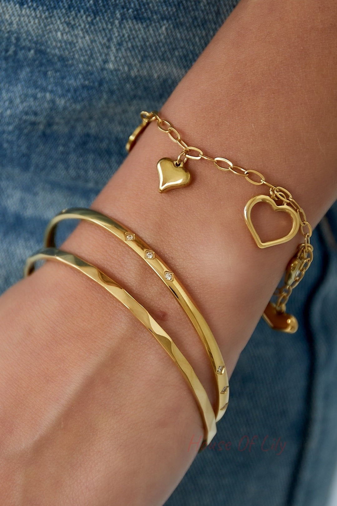 Wrist wearing layered bracelets including a textured gold-tone bangle, diamond-studded slim bangle, and a chain bracelet with gold heart charms