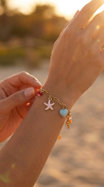 Coastal Charm gold link bracelet featuring pink starfish, light blue heart, and colorful bead charms on a sunny beach.