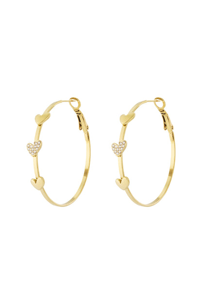 Gold hoop earrings decorated with three heart-shaped charms, one encrusted with small crystals, displayed on white background