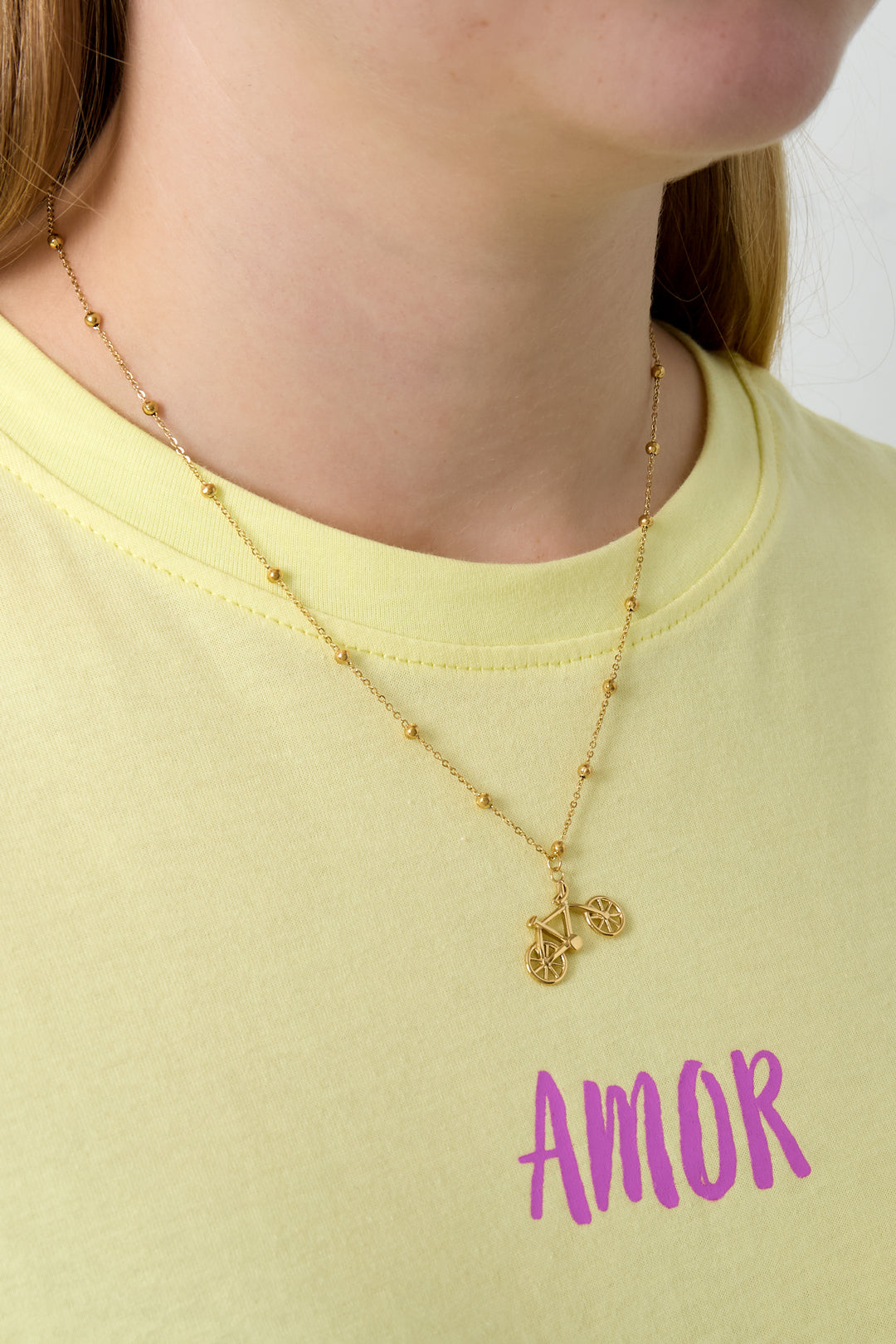 Close-up of woman wearing gold bicycle pendant necklace over yellow shirt with Amor text