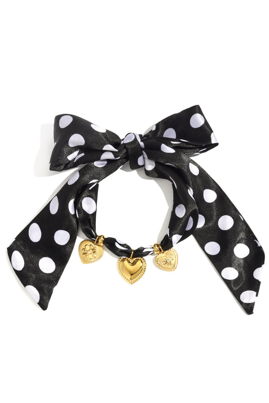 Black polka dot fabric bow bracelet with three gold heart-shaped charms against white background