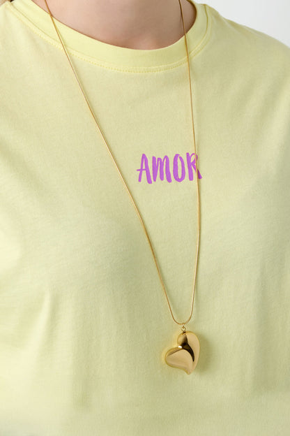 Gold heart pendant necklace worn over a pastel yellow t-shirt with the word 'AMOR' printed in purple