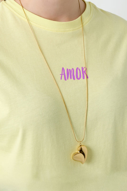 Gold heart pendant necklace worn over a pastel yellow t-shirt with the word 'AMOR' printed in purple