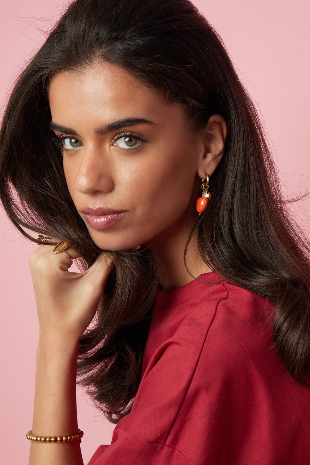 A woman wears Coral Crush Heart Hoops: gold-toned huggie earrings featuring a small, vibrant orange heart charm.