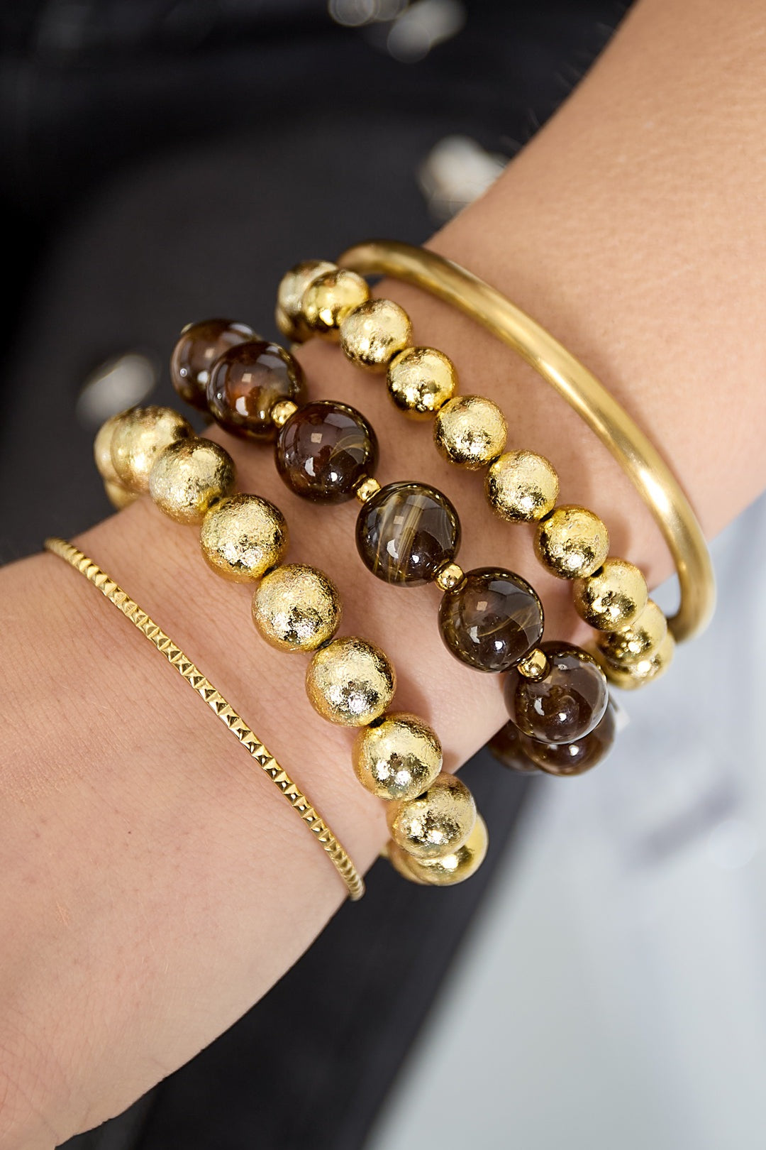 Close-up of wrist with bracelets including dark brown marbled and textured gold bead bracelets styled with gold bangle