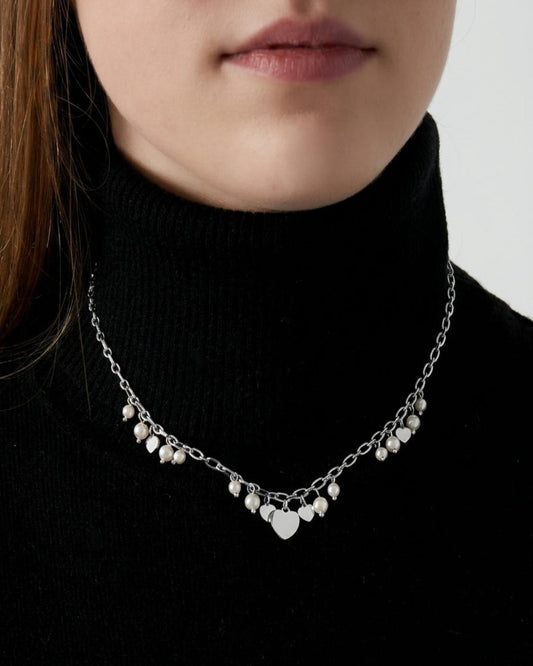 A close-up of a model wearing a delicate silver chain necklace with small pearl-like beads and a central heart-shaped charm, styled with a black turtleneck sweater.