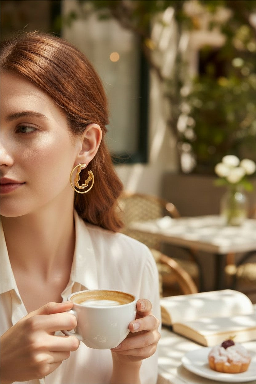 Petit Coeur Hoops: gold, open-circle earrings featuring a unique, wavy interior detail, worn by a woman at a cafe.